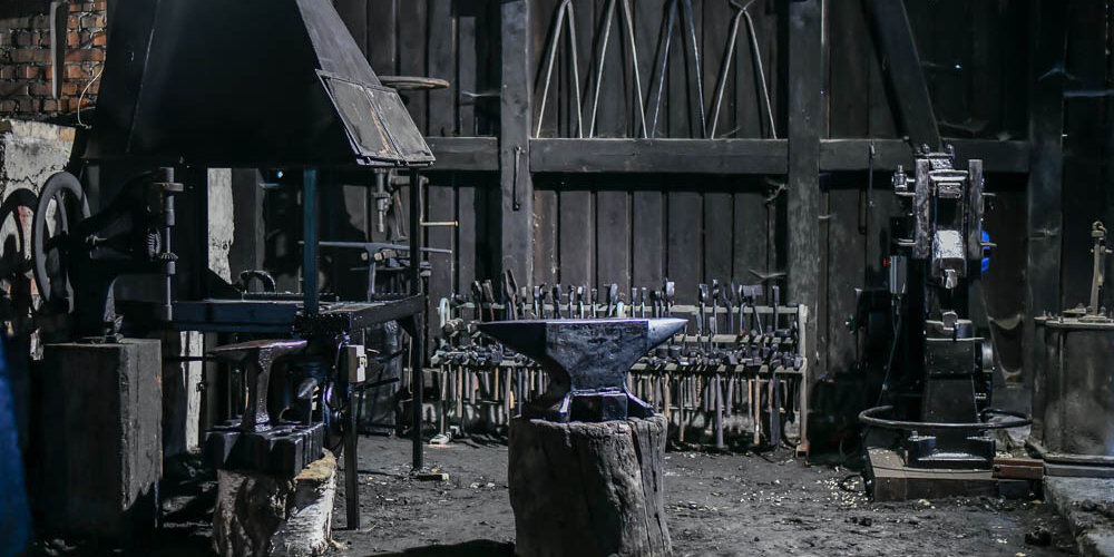 Zdjęcie przedstawia wnętrze Kuźni Wodnej w Oliwie. Na pierwszym planie po lewej okap paleniska, metalowe kowadło i przyrząd. Za nimi zawieszone na drewnianej konstrukcji metalowe narzędzia.  