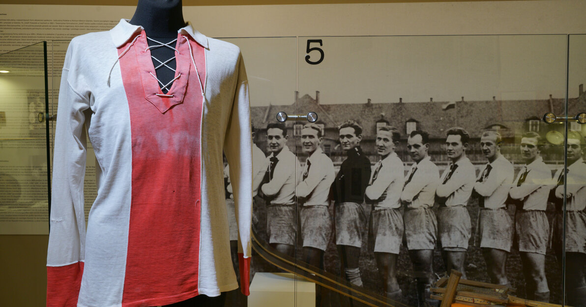 Zdjęcie przedstawia koszulkę piłkarską KS Gedania z długimi rękawami. Środek jest czerwony, boki i rękawy są białe. W tle czarno-białe zdjęcie przedstawiające sportowców z dwudziestolecia międzywojennego ustawionych w szeregu spoglądających na widza.   
