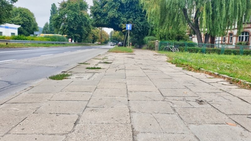 Zdjęcie przedstawia chodnik oraz część jezdni. W tle widać drzewa oraz budynek