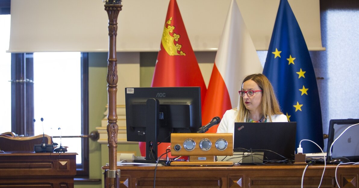 Na zdjęciu znajduje się przewodnicząca Rady Miasta, która siedzi przed monitorem komputera, trwają zdalne obrady Rady, za kobietą znajdują się flagi Gdańska, Polski i Unii Europejskiej. W tle widać, też opuszczony ekran do wyświetlania projekcji multimedialnych.