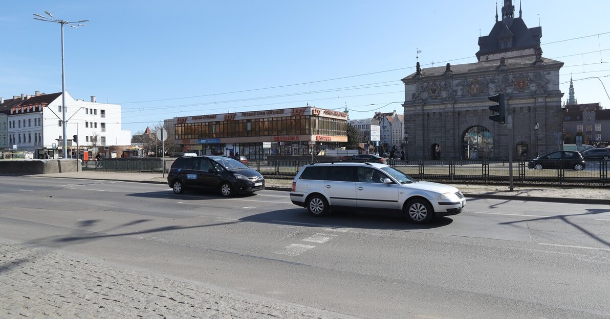 Zdjęcie przestawia ulicę wraz z jadącymi samochodami. Za ulica widać Bramę Wyżynną oraz budynek LOT.
