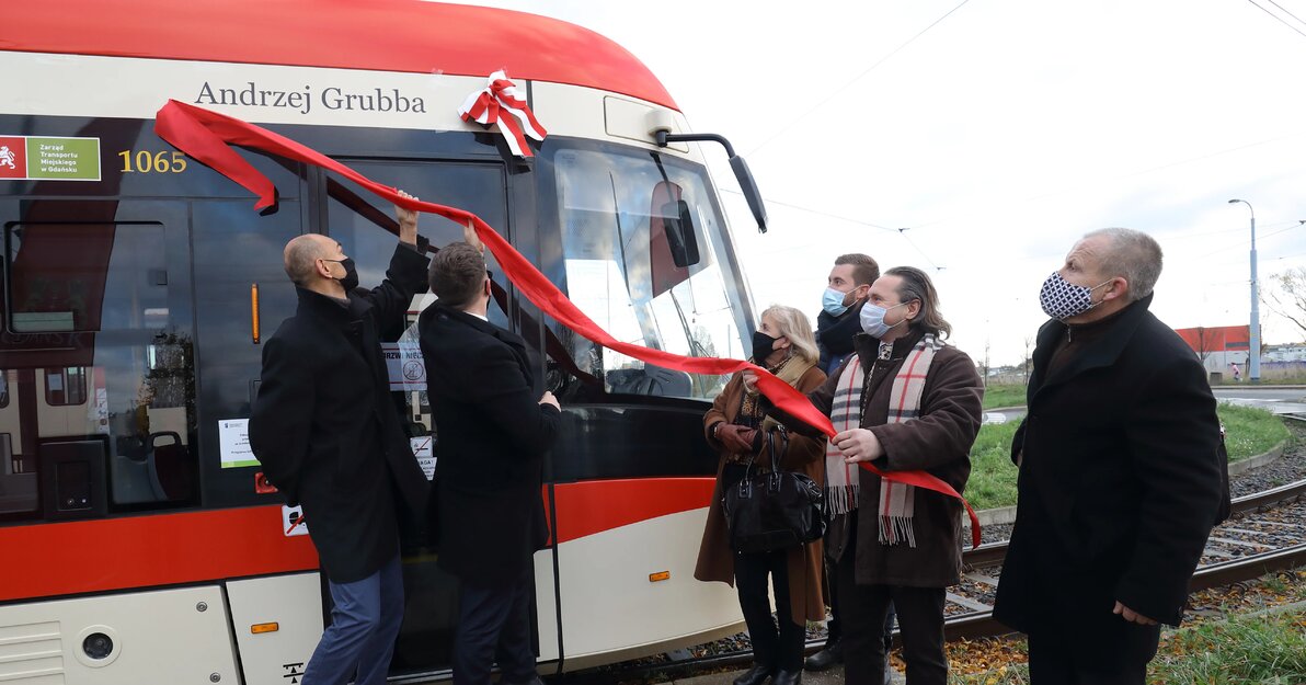 Uczestnicy uroczystości ściągają z burty tramwaju czerwoną wstęgę, spod której wyłania się napis: Andrzej Grubba. Po lewej stronie stoją Leszek Kucharski i Piotr Borawski. Po prawej stronie Lucyna Grubba z synem oraz Mateusz Lipowski i Włodzimierz Machnikowski. 