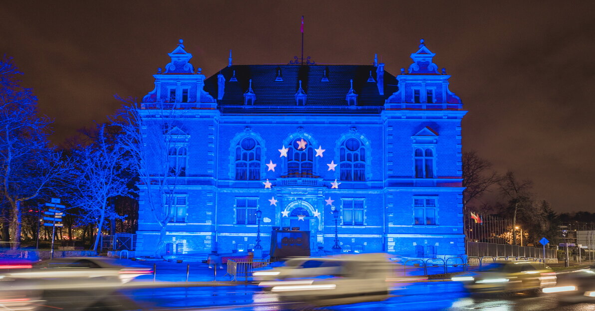 Zdjęcie przedstawia budynek, siedzibę Rady Miasta intensywnie podświetloną na niebieski kolor. Na pierwszym planie widać także ulicę i sfotografowane w pędzie, rozmazane samochody. Na mokrej od deszczu jezdni odbijają się światła. 