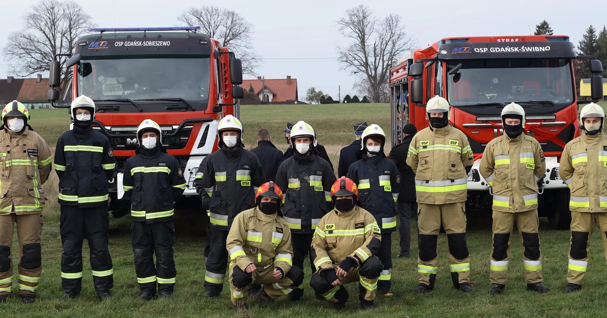 Na zdjęciu dwa nowe wozy strażackie, przed nimi stoją dwa zastępy strażaków z Ochotniczej Straży Pożarnej.