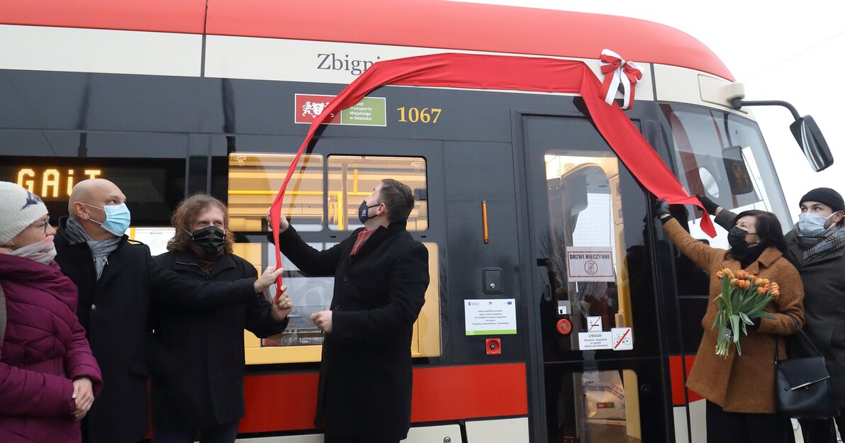 Uczestnicy uroczystości nadania tramwajowi imienia Zbigniewa i Macieja Kosycarzy odsłaniają napis z imionami i nazwiskiem patronów. Napis wyłania się spod czerwonej wstęgi. 