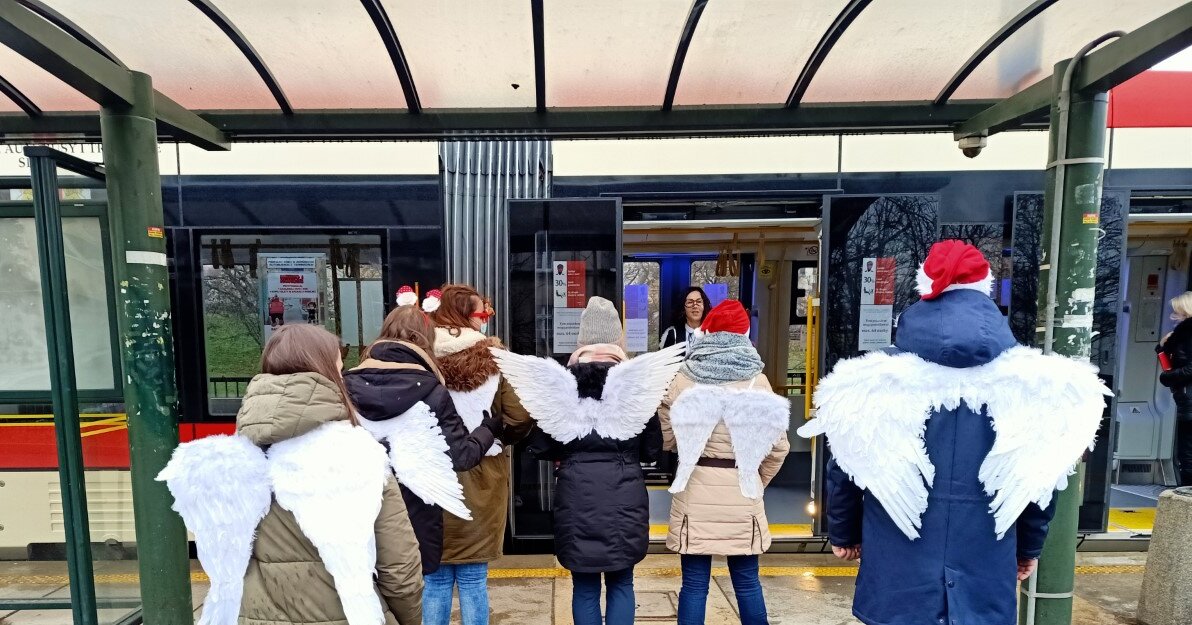 Osoby stoją na przystanku i czekają na wejście do tramwaju. Sfotografowani są od tyłu. Na plecach mają białe skrzydła. Dwie osoby mają na głowach czapki Mikołaja. 