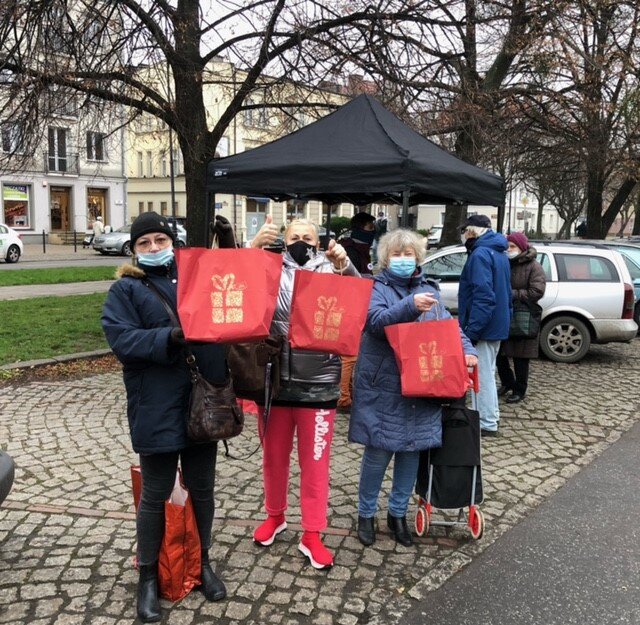 Społeczni opiekunowie kotów prezentują pakiety z karmą dla wolnobytujących kotów. W tle namiot, pod którym wydawane są pakiety. 