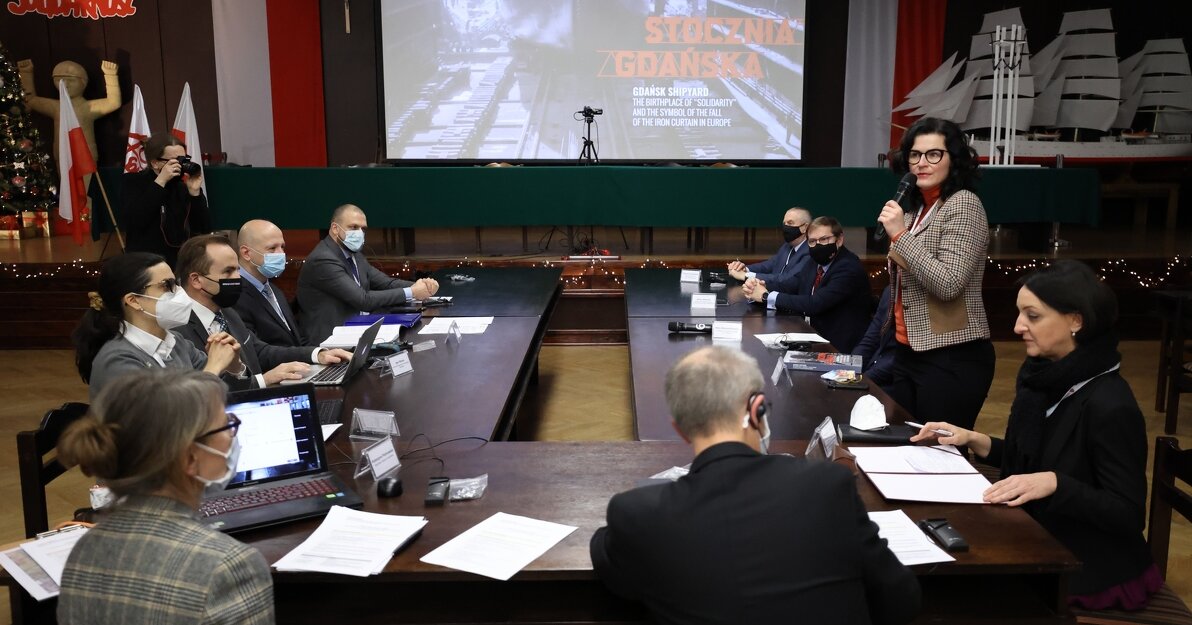 Przy stole zasiadają przedstawiciele różnych organizacji. Przemawia na stojąco Aleksandra Dulkiewicz, w tle ekran z czołową storna prezentacji o Stoczni Gdańskiej. 