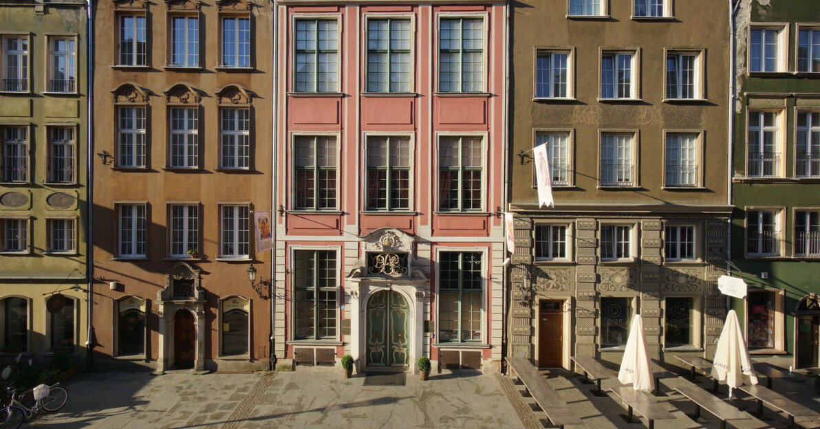 Zdjęcie przedstawia fragment ulicy Długiej na tle błękitnego nieba. W centrum trzy kamiennice. W środku wyróżniająca się, jasnoczerwona fasada kamienicy Uphagenów przy Długiej 12 z licznymi oknami. 