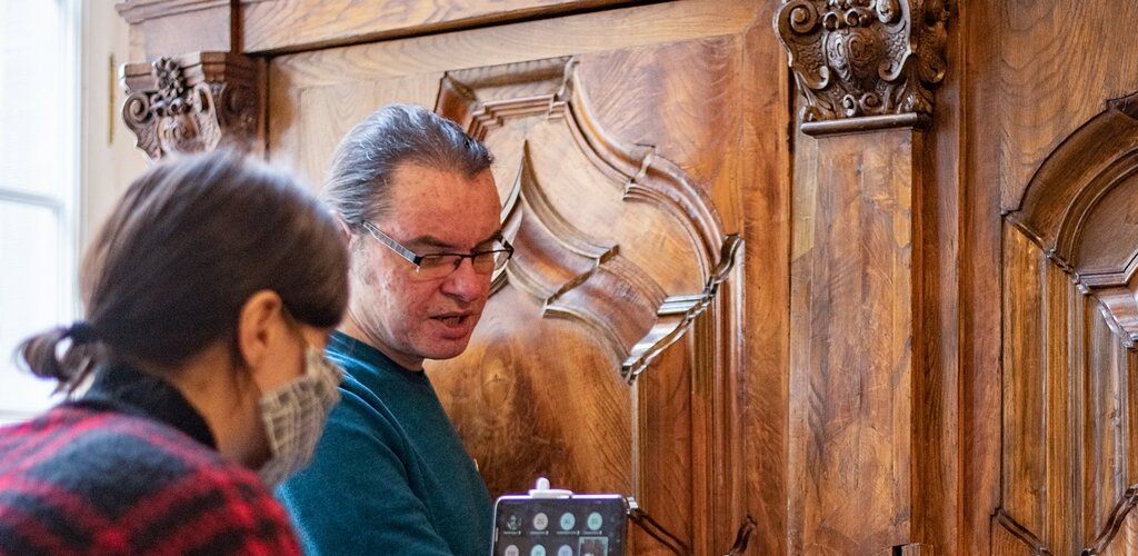 Zdjęcie przedstawia dwie osoby. Kobieta trzyma kijek, a na nim telefon. Po lewej długowłosy mężczyzna w okularach wskazuje na element starej, masywnej szafy i opowiada o nim. Fot. A. Grabowska (Muzeum Gdańska) 