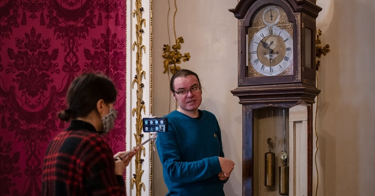 Zdjęcie przedstawia dwie osoby. Kobieta trzyma kijek, a na nim telefon. Po lewej w głębi długowłosy mężczyzna w okularach patrzy w obiektyw i opowiada do kamery o starym zabytkowym zegarze. 