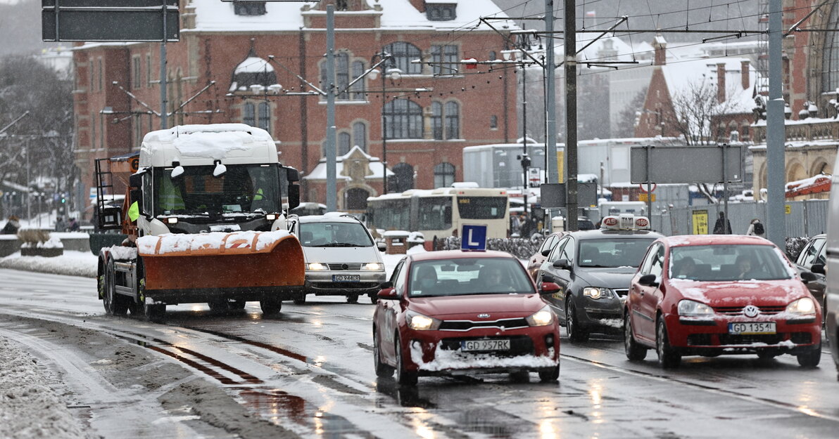 Ulica po której jadą samochody, wśród nich jest też pojazd pługoposypywarki. Na drodze i chodniku zalegają duże zaspy śniegu