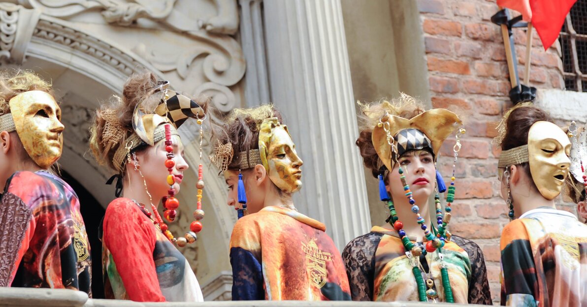 Na zdjęciu zrobionym z dołu znajduje się pięć kobiet ubranych w kolorowe stroje. Do ozdób na ich głowach są przymocowane długie barwne korale. Trzy z nich są odwrócone plecami do widza, na tyłach głów mają założone złote maski przedstawiające twarze. 