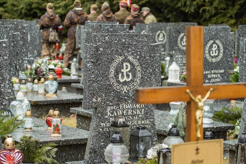 Zdjęcie kolorowe. W kadrze na pierwszym planie po prawej drewniany krzyż, za nim kamienne nagrobki. W centrum nagrobek porucznika Edwarda Kawerskiego. Nad napisem symbol Polski Walczącej otoczony laurem. W tle rozmyte sylwetki harcerzy.