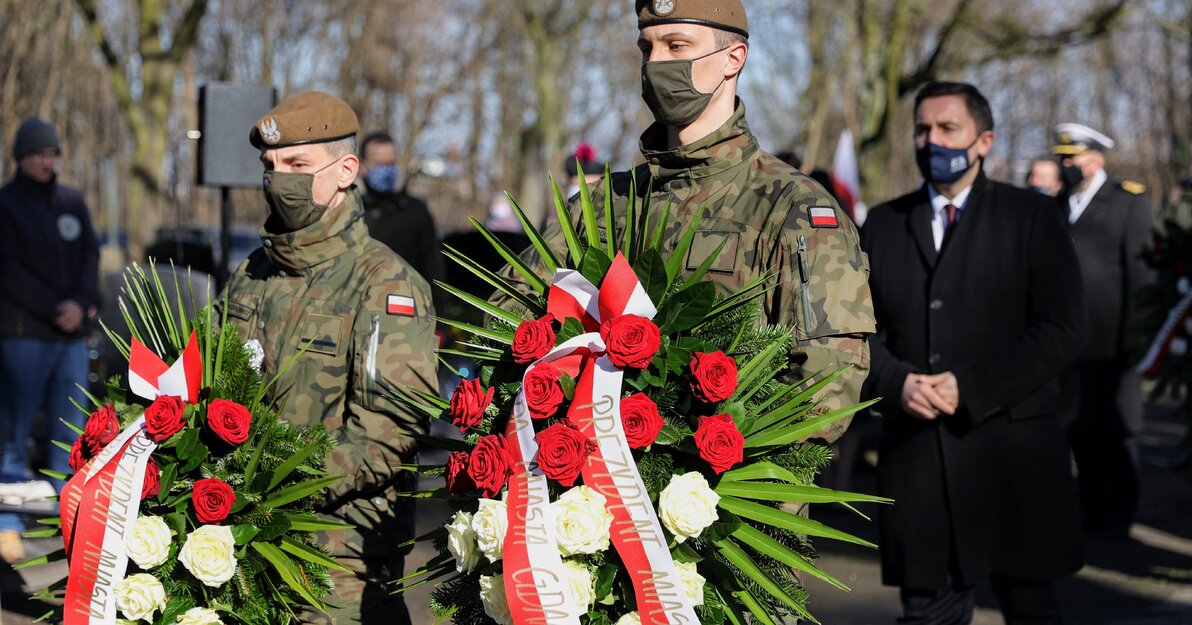 Zdjęcie przedstawia dwóch żołnierzy niosących wieńce biało-czerwonych róż. Za nimi idzie Piotr Borawski, zastępca prezydenta. Na szarfach wieńców widać napisy "Prezydent Miasta Gdańska i Rada Miasta Gdańska". 