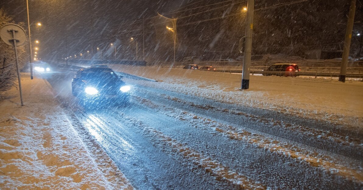 Zaśnieżona ulica, jadący po niej samochód i silnie padający śnieg.