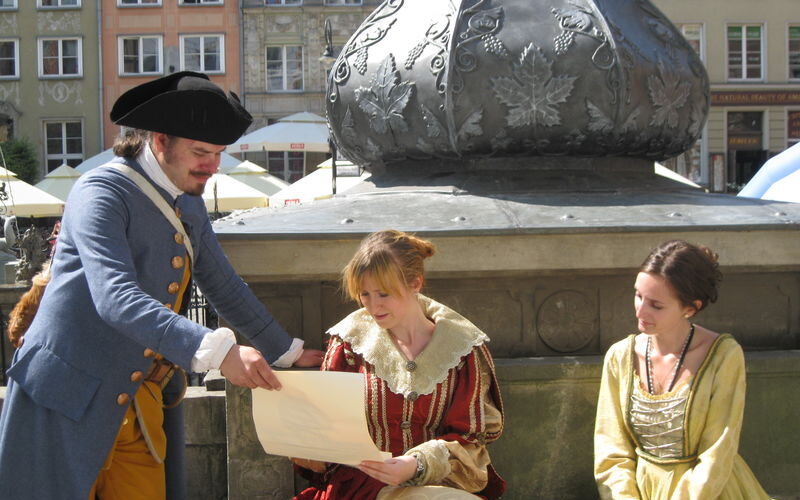 Zdjęcie. Przestrzeń. 3 postacie przebrane w stroje dawnych mieszczan na przedprożu Dworu Artusa w Gdańsku. Z lewej stoi mężczyzna w trójkątnym kapeluszu, po prawej dwie mieszczanki. Czytają trzymany przez mężczyznę w dłoni list. 