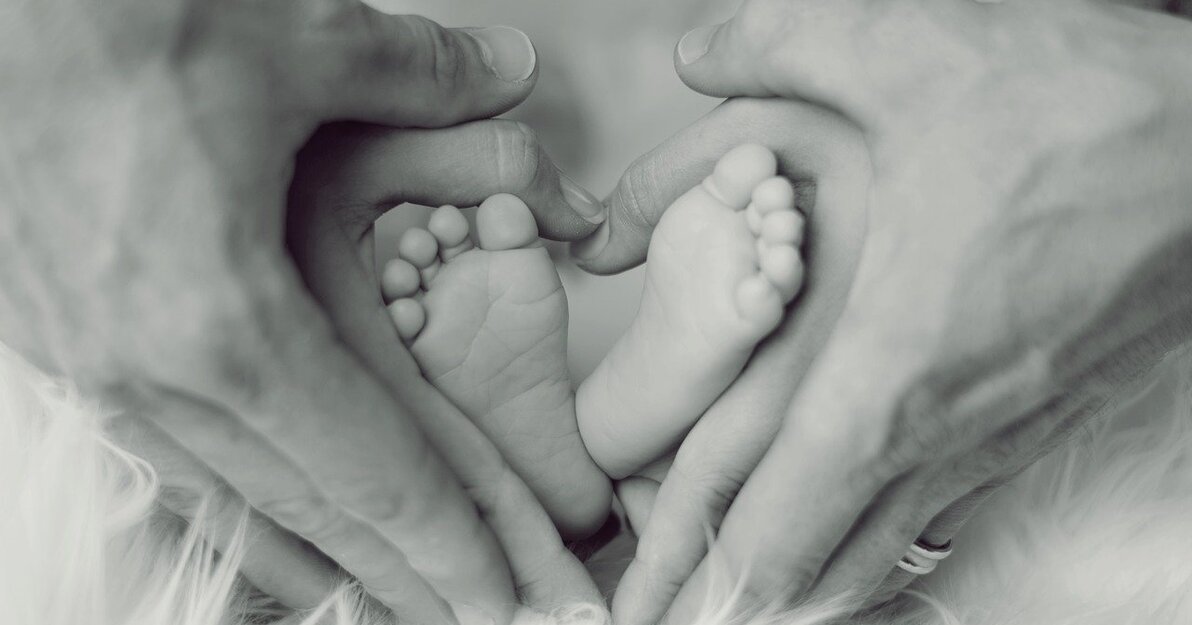 Na zdjęciu dłonie kobieca i męska, które obejmują stópki noworodka