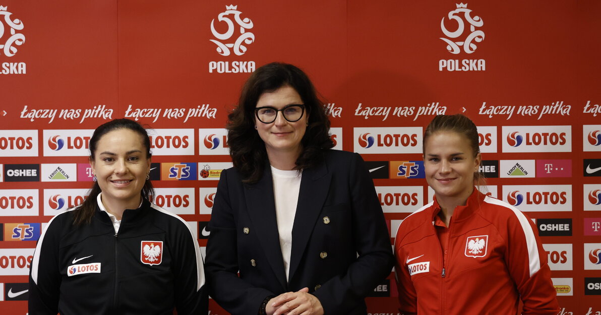 Aleksandra Dulkiewicz, prezydent Gdańska z zawodniczkami kobiecej reprezentacji Polski w piłce nożnej. Za paniami czerwone tło z logotypami. 