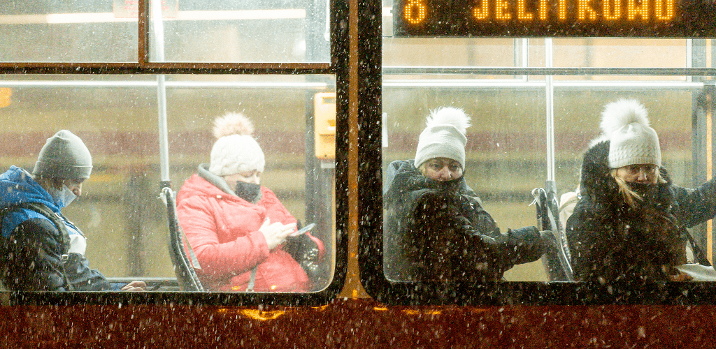 Zdjęcie. Fragment tramwaju z dwoma oknami. Wewnątrz siedzą 4 osoby z maseczkami zakrywającymi częściowo twarz. Patrzą  w telefon, albo w szybę (w kierunki widza). W prawym oknie u góry napis "8 Jelitkowo". Pada śnieg.   