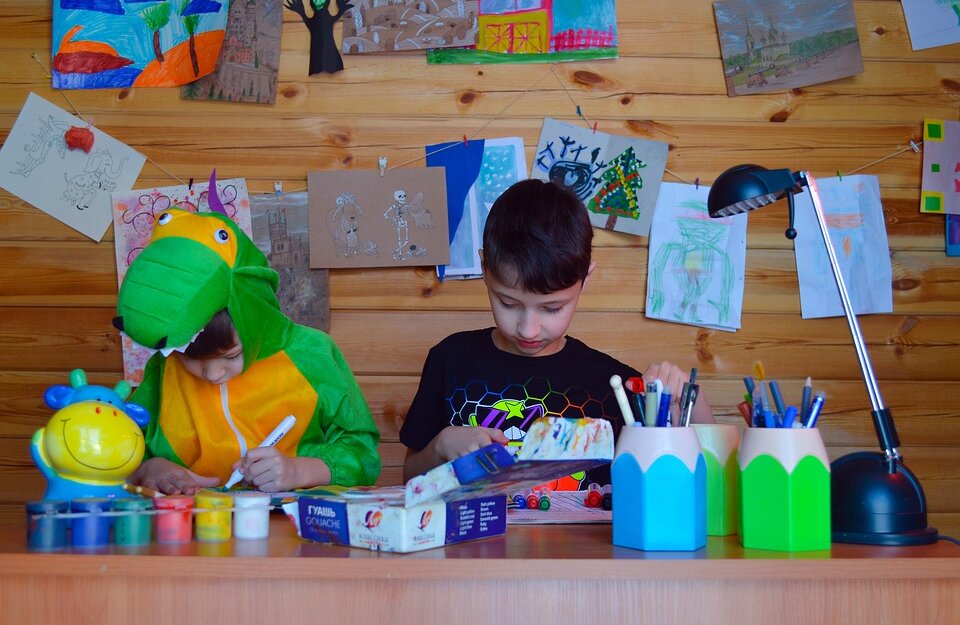 Zdjęcie przedstawia chłopców podczas wykonywania prac plastyczny. Jeden z nich jest ubrany w strój dinozaura. Na stole znajdują się kredki i farby. Za chłopcami na ścianie widzą rysunki. 