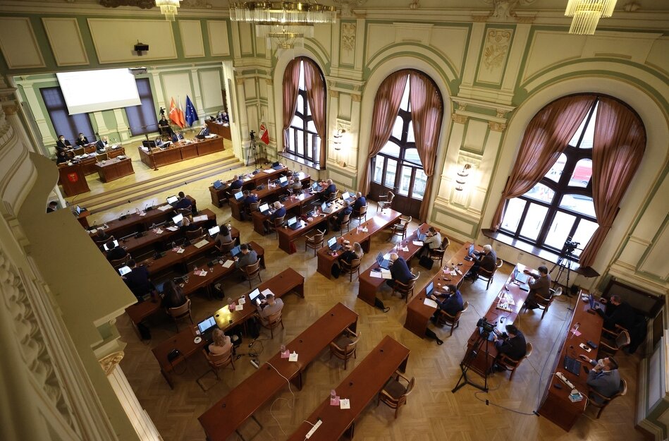 Sala obrad Rady Miasta. Na pierwszym planie biurka radnych z mikrofonami i przyciskami do głosowania, na drugim planie dwa okna Sali Obrad.