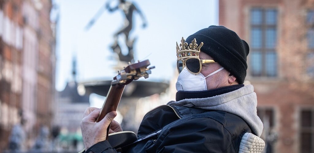 Zdjęcie. Mężczyzna z maseczką na twarzy i koroną na głowie gra na gitarze. W tle Fontanna Neptuna. 