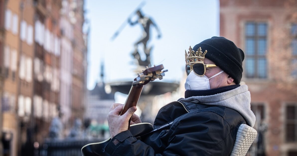 Zdjęcie. Mężczyzna z maseczką na twarzy i koroną na głowie gra na gitarze. W tle Fontanna Neptuna. 