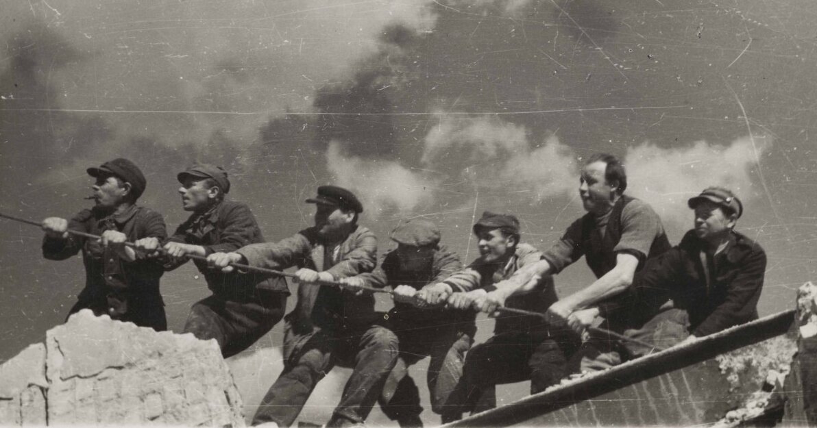 Zdjęcie przedstawia grupę mężczyzn trzymających naprężoną linę na dachu budynku. 