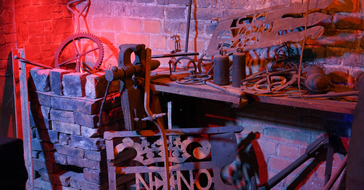 Zdjęcie przedstawia aranżację warsztatu kowalskiego. Na półkach leżą liczne narzędzia, np. obcęgi, młoty i szczypce.