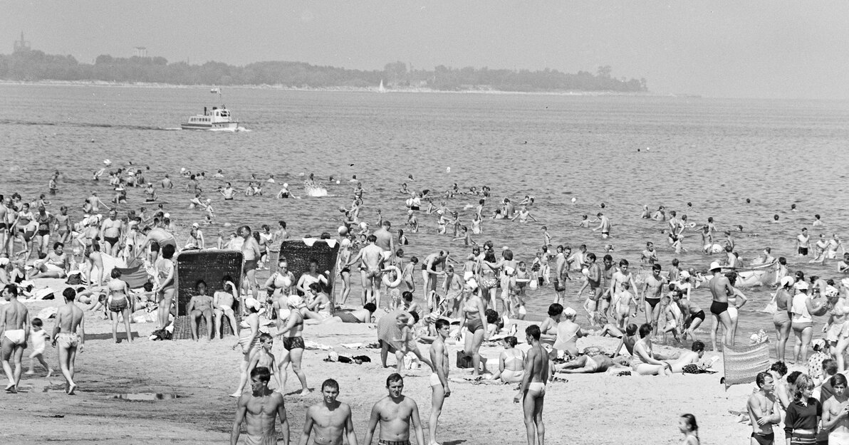 Fragment czarno-białej fotografii. Na zdjęciu plaża na gdańskich Stogach. Na brzegu i w wodzie liczni ludzie. W tle fragment brzegowy w kierunku Jelitkowa. Na morzu niewielki statek wycieczkowy. 