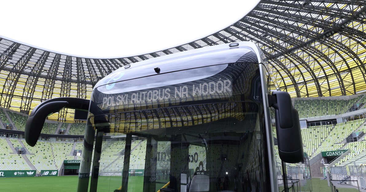 Autobus stoi na płycie stadionu. Ujecie od przodu. Na wyświetlaczu napis: polski autobus na wodór. W tle murawa i trybuny Polsat Plus Arena Gdańsk.  