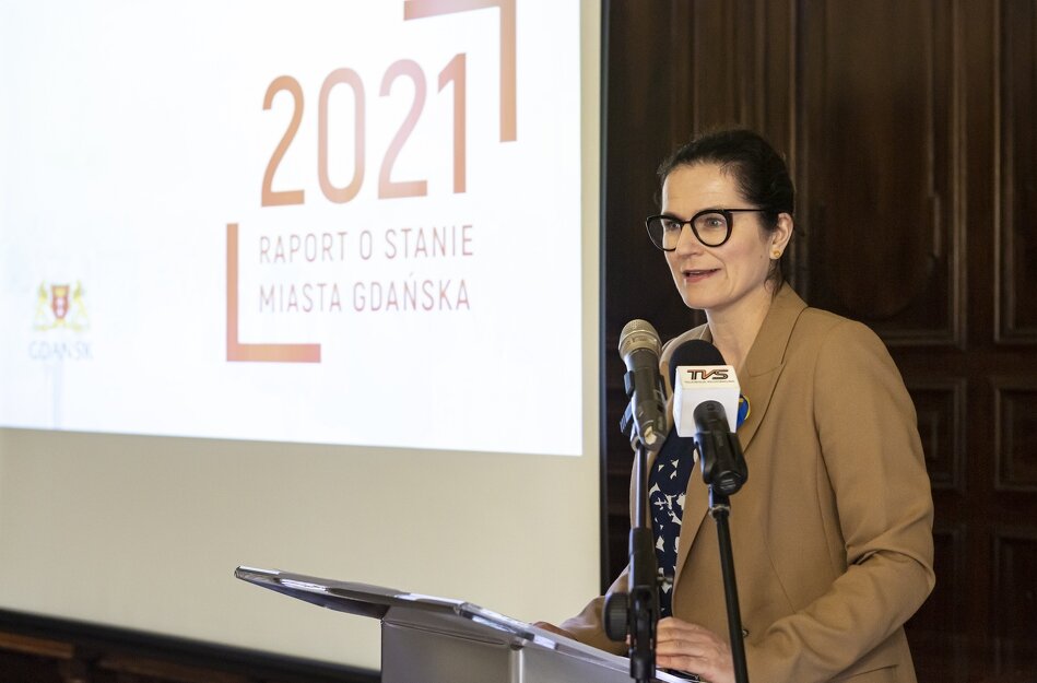 Prezydent Aleksandra Dulkiewicz na mównicy. Za nią, po lewej stronie, wyświetlony tytułowy slajd z Raportem o stanie miasta za rok 2021.