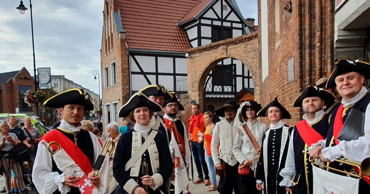 Zdjęcie przedstawia stojące w dwóch rzędach grupkę ludzi przebranych w historyczne stroje. W tle fragment fasady kościoła św. Katarzyny oraz Małego Młyna.  