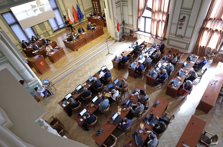 Sala obrad Rady Miasta Gdańska widziana z góry. Większą część zdjęcia zajmują radni siedzący za stołami, przed nimi laptopy. Z prawej widoczne dwa duże okna. Z lewej, na podwyższeniu, prezydium rady, nad którym zawieszony jest duży ekran.