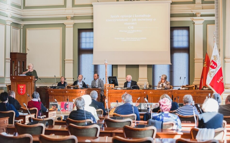 Obrady wrześniowej sesji Rady Seniorów. Widok na Salę obrad od tyłu, w stronę prezydium. Na pierwszym planie radni za pulpitami, w głębi prezydium i telebim.