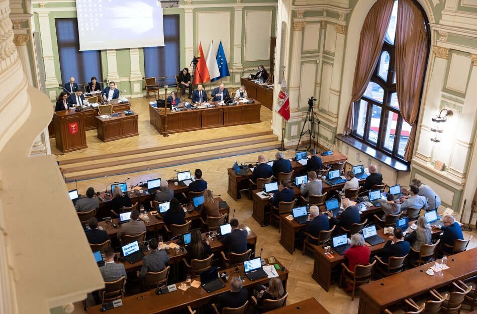 Sala obrad Rady Miasta Gdańska widziana z góry. Większą część zdjęcia zajmują radni siedzący za stołami, przed nimi laptopy. Z prawej widoczne dwa duże okna. Z lewej, na podwyższeniu, prezydium rady, nad którym zawieszony jest duży ekran. 