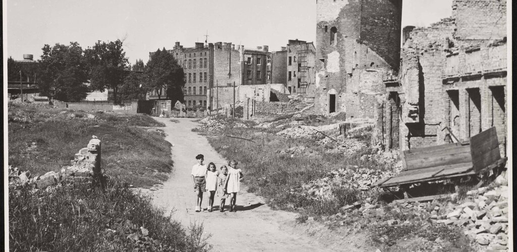 Fotografia czarno-biała ukazuje widok na zniszczone w wyniku II wojny światowej zabudowania Gdańska - okolice Targu Rybnego. Na pierwszyn planie droga, po której idzie czwórka dzieci. Po prawej stronie widoczna baszta Łabędź, w oddali zabudowania ulicy Grodzkiej.
