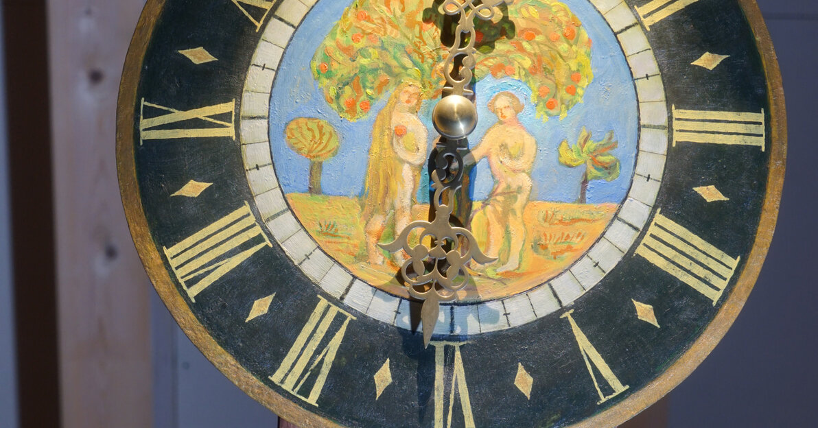 Zdjęcie przedstawia tarczę zegara żuławskiego. Na okrągłej tarczy namalowano w kolorze scenę z biblijnego raju – Ewę i Adama stojących pod drzewem. Rysunek otacza czarna obwódka, na której umieszczonych jest dwanaście cyfr rzymskich wskazujących godziny. 