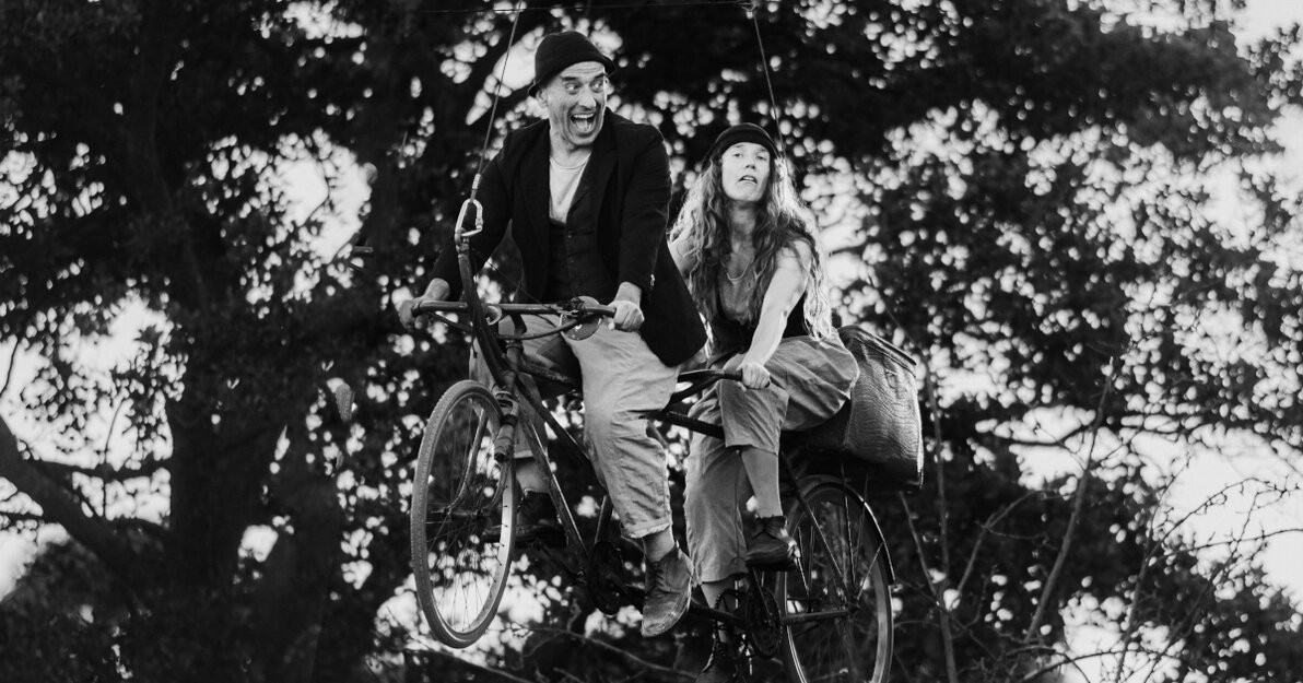 Zdjęcie czarno białe. Spektakl uliczny. Mężczyzna i kobieta siedzą na rowerze dla dwóch osób, tandemie. Rower unosi się w powietrzu, zawieszony na linkach.