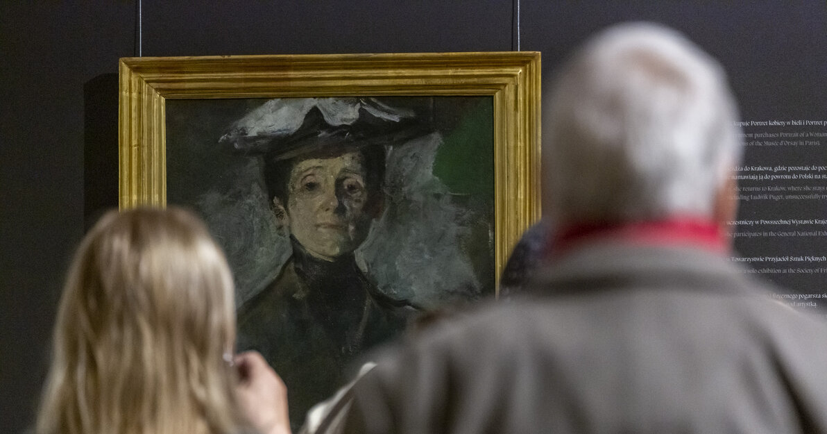 Wernisaż wystawy Olgi Boznańskiej. Zdjęcie przedstawia dwie osoby, mężczyznę i kobietę (po prawej) stojących plecami. Za nimi w tle fragment autoportretu artystki. 