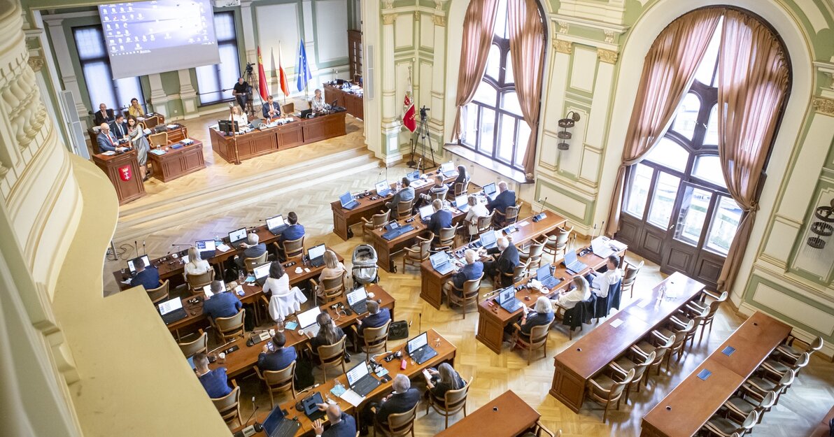 Sala obrad Rady Miasta Gdańska widziana z góry. Większą część zdjęcia zajmują radni siedzący za stołami, przed nimi laptopy. Z prawej widoczne dwa duże okna. Z lewej, na podwyższeniu, prezydium rady, nad którym zawieszony jest duży ekran. 