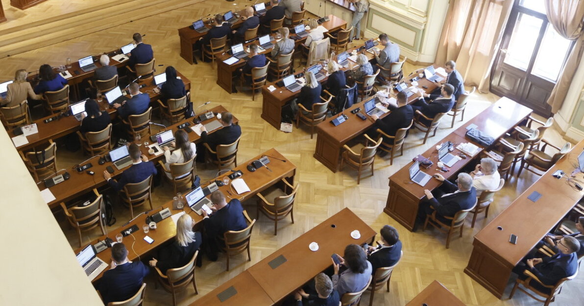 Sala obrad Rady Miasta Gdańska widziana z góry. Większą część zdjęcia zajmują radni siedzący za stołami, przed nimi laptopy. Z prawej widoczne dwa duże okna. Z lewej, na podwyższeniu, prezydium rady, nad którym zawieszony jest duży ekran. 