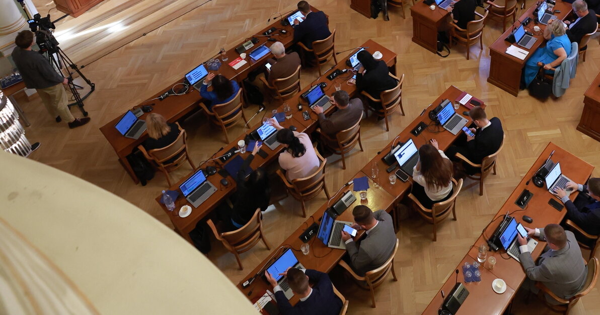 Sala obrad Rady Miasta Gdańska widziana z góry. Większą część zdjęcia zajmują radni siedzący za stołami, przed nimi laptopy. Z prawej widoczne dwa duże okna. Z lewej, na podwyższeniu, prezydium rady, nad którym zawieszony jest duży ekran. 