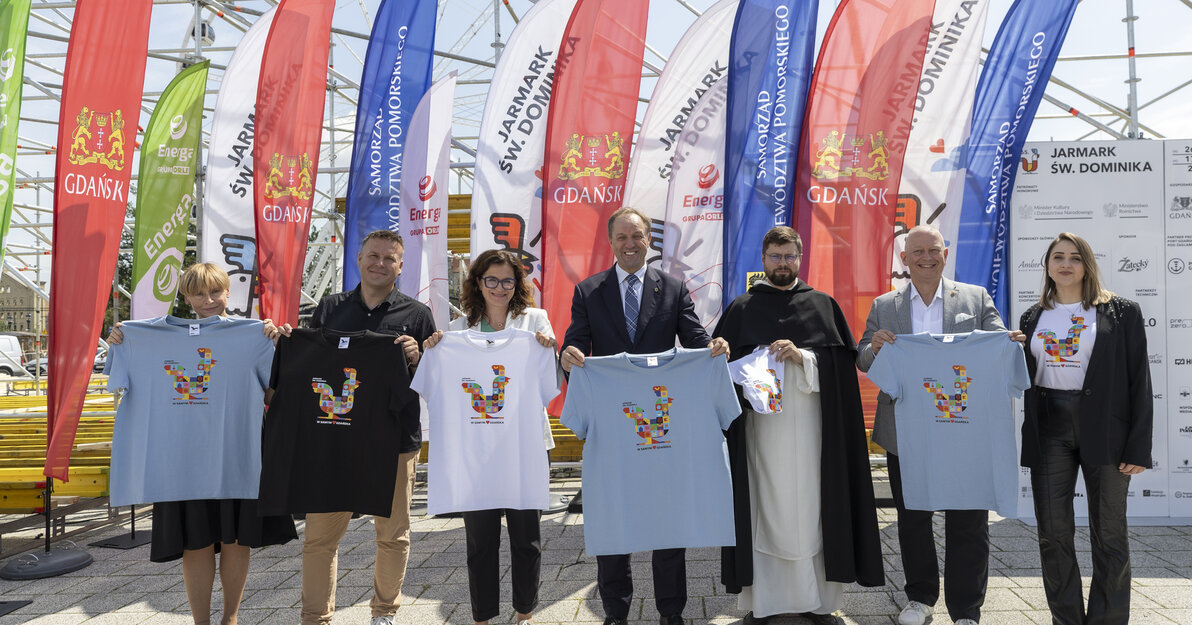 Sześć osób stoi w szeregu, trzymając przed sobą koszulki z kolorowym nadrukiem przedstawiającym jarmarkowe koguty. W tle powiewają wysokie, wielobarwne flagi z napisami „JARMARK ŚW. DOMINIKA” i „GDAŃSK”. Po prawej stronie znajduje się tablica z logotypami sponsorów wydarzenia.