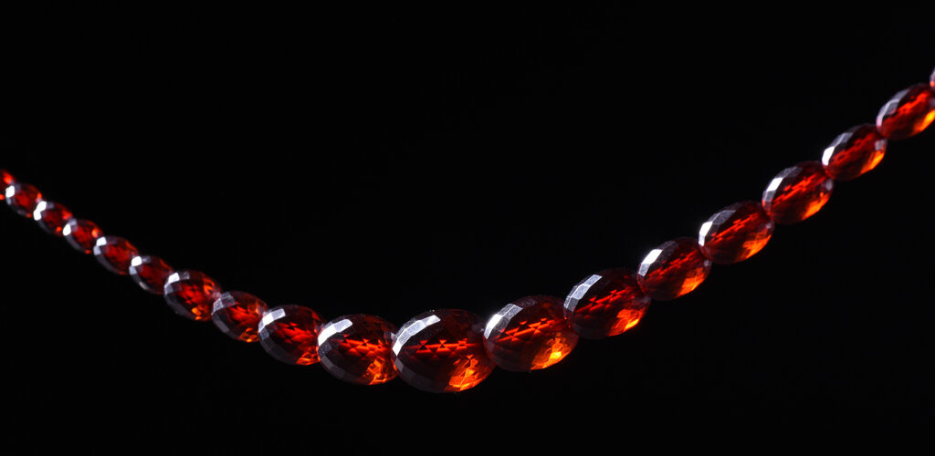 Sznur korali fasetowanych. Paciorki owalne o różnej wielkości, zwiększające się od zapięcia do środka sznura. Zapięcie plastikowe, dwuczęściowe, zamykane przez zakręcanie. Paciorki przejrzyste w kolorze koniakowym - imitacja bursztynu.