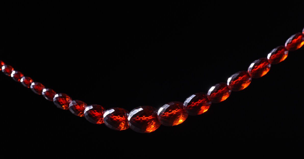 Sznur korali fasetowanych. Paciorki owalne o różnej wielkości, zwiększające się od zapięcia do środka sznura. Zapięcie plastikowe, dwuczęściowe, zamykane przez zakręcanie. Paciorki przejrzyste w kolorze koniakowym - imitacja bursztynu.