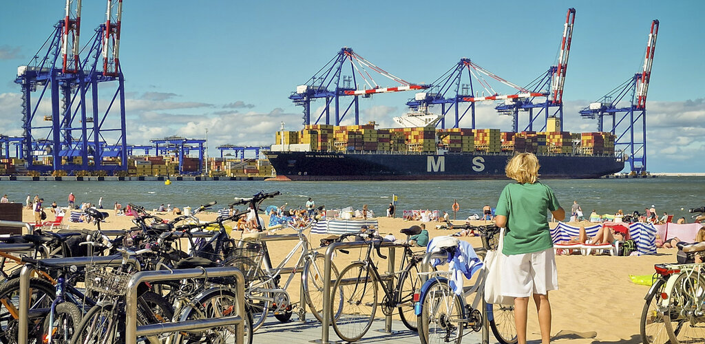 Kobieta stoi boso na plaży obok zaparkowanych rowerów i patrzy na ogromny kontenerowiec oraz suwnice portowe; plażowicze wypoczywają na piasku.
