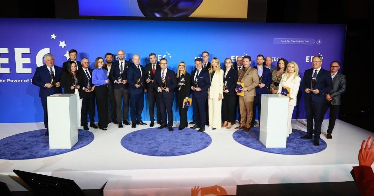 Na szerokiej scenie stoi kilkanaście osób w formalnych strojach, ustawionych w rzędzie i trzymających w dłoniach szklane statuetki. Tło jest niebieskie, z dużymi napisami „EEC” i „Top Inwestycje 2026”, a scena oświetlona jasnym światłem. Całość ma uroczysty, oficjalny charakter wręczenia nagród.